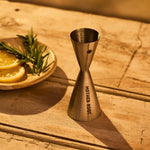 A silver metal shot glass with a sun emblem, branded with 'MOTHER ROOT', placed on a wooden surface next to a plate with lemon slices and a sprig of rosemary.