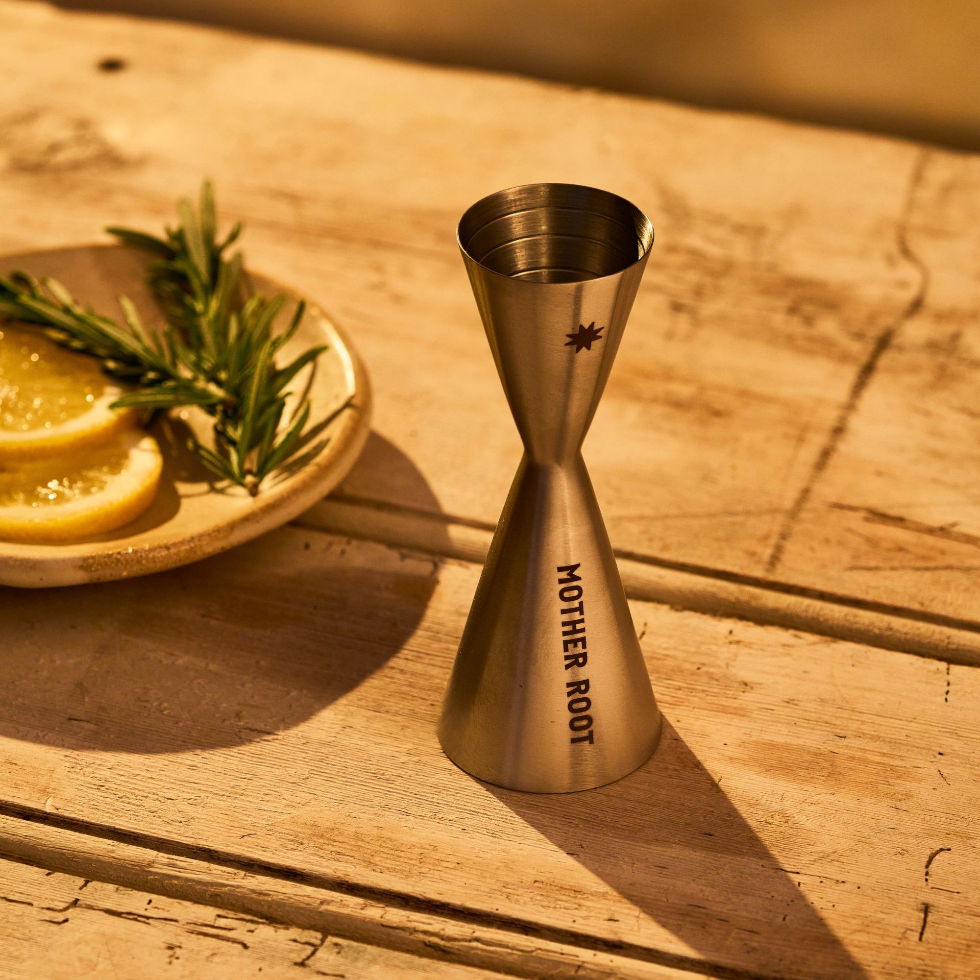 A silver metal shot glass with a sun emblem, branded with 'MOTHER ROOT', placed on a wooden surface next to a plate with lemon slices and a sprig of rosemary.